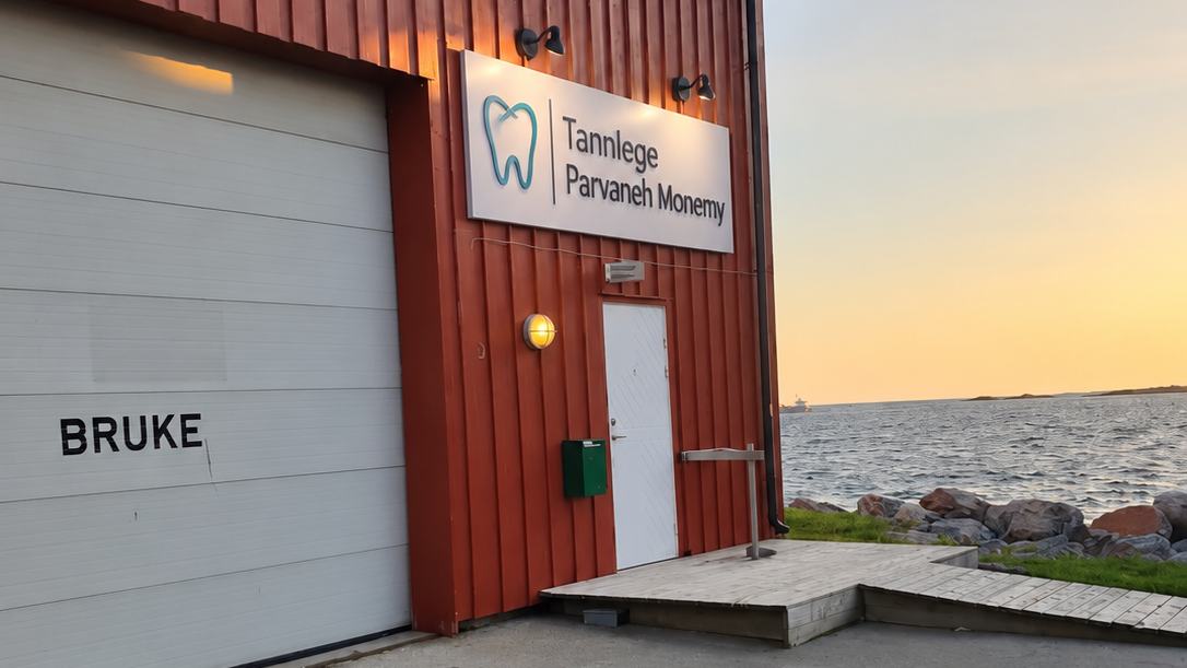 Inngangen til tannklinikken: hvit dør under skiltet «Tannlege Parvaneh Monemy» på det røde bygget ved sjøen på Sistranda